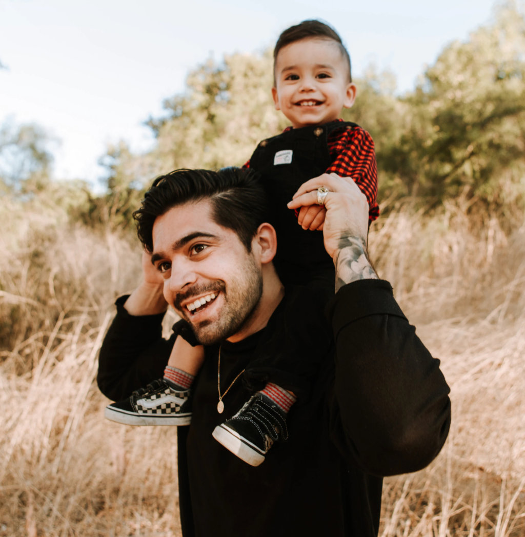Latino father smiling while carrying his infant son on his shoulders in a sunny park.