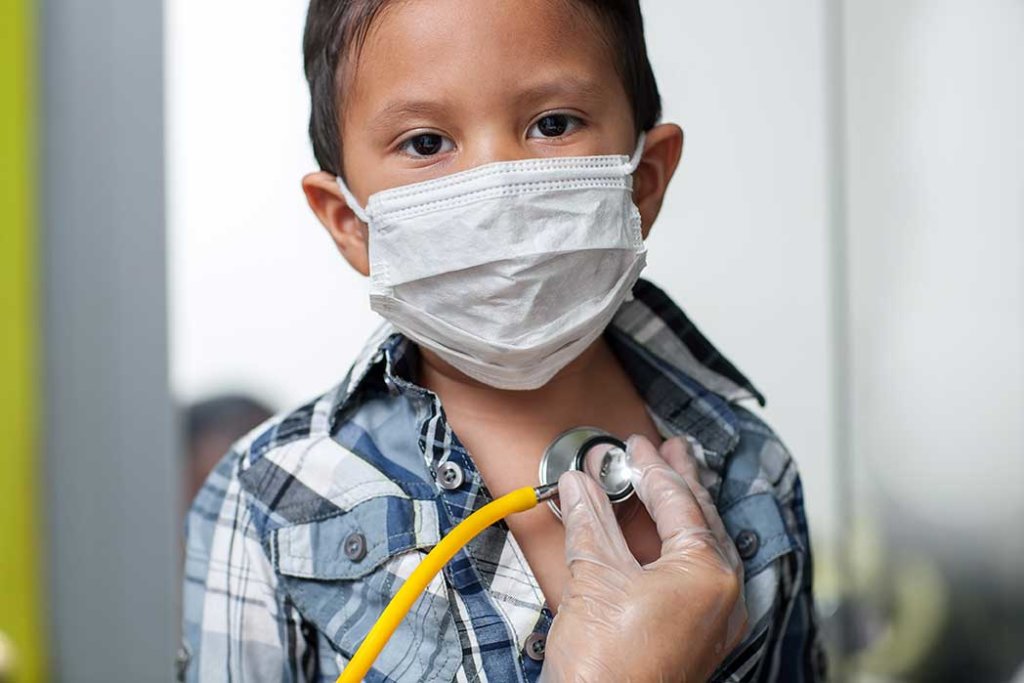 Child wearing a medical mask to prevent spread of virus is getting a heart screening with stethoscope placed on chest.