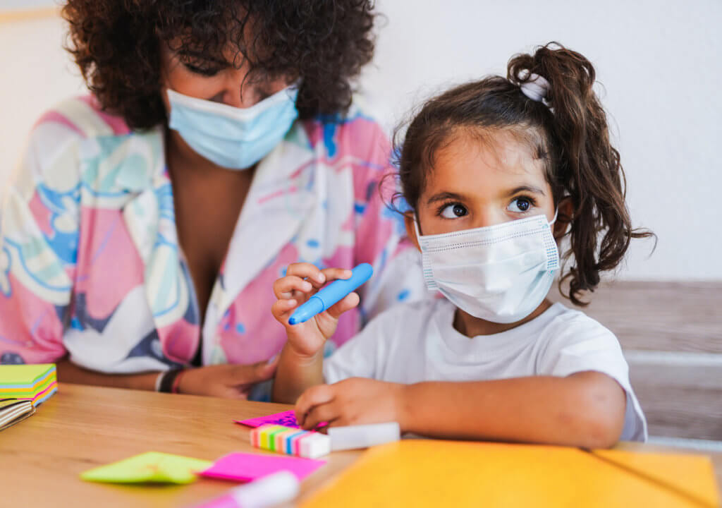 Teacher draws with little girl while wearing surgical face mask - Safety measures | Latino children and health