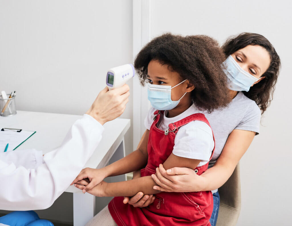 Pediatrician checking body temperature of a child using a non-contact thermometer. African American girl with her mom wearing protective masks at the doctor's consultation