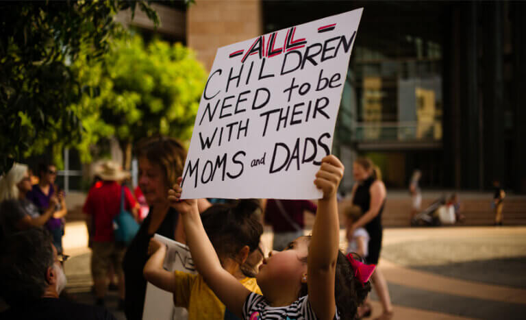 Little girl holding sign stating "All Children Need to be with Their Moms and Dads."