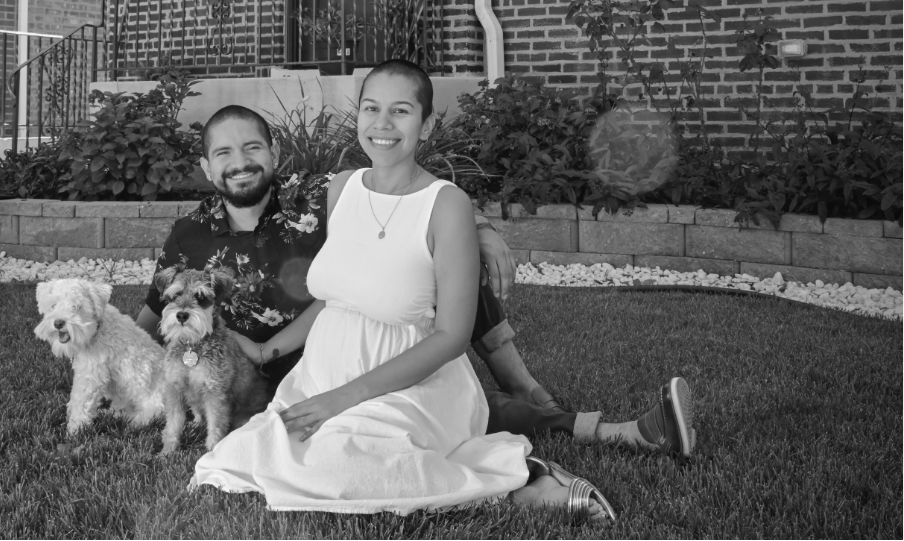 A black and white photo of a couple sitting on the grass in front of a brick house with two hairy dogs.