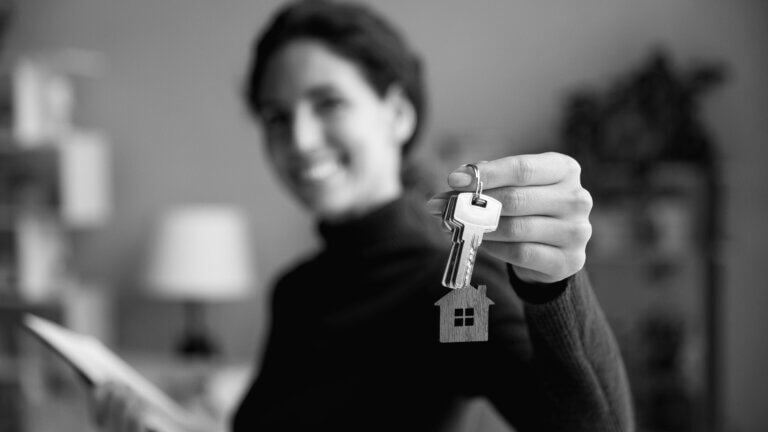 A black and white photograph of a woman, out of focus, holding a set of keys with a house shaped keychain, in focus, in front of the camera.