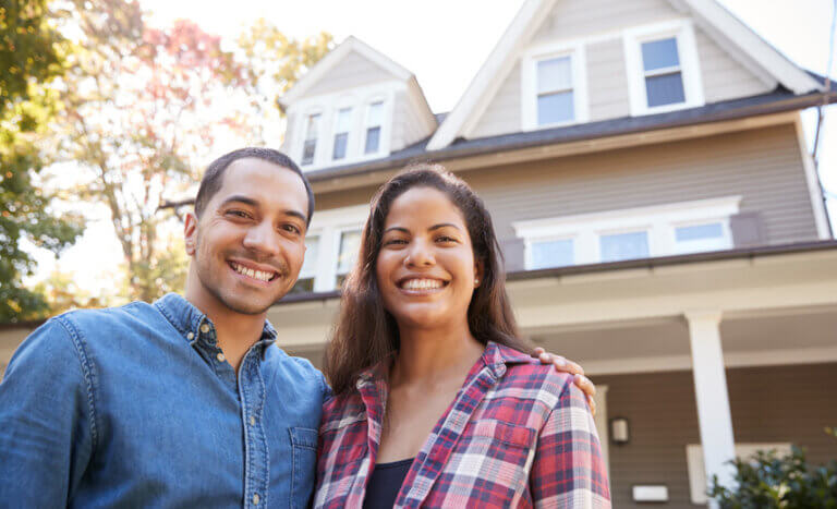 Latino couple outside of their newly purchased home smiling for the camera.