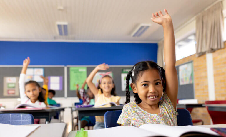 Elementary school aged children raising their hands in a classroom waiting to be picked to answer a question.