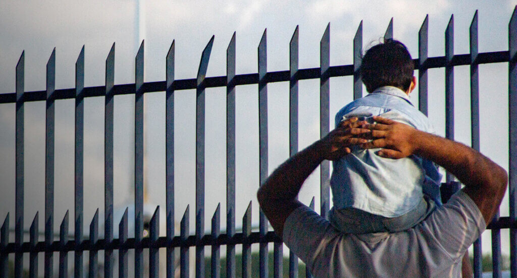 Man with child on his shoulders looking at border wall fence