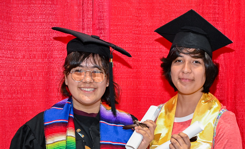 Two young Latinas pose with diplomas while wearing valedictorian caps and gowns.